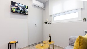 A modern, well-lit room with a TV mounted on the wall, a small table with a bottle and glasses, a sofa with a cushion, and a window providing natural light.