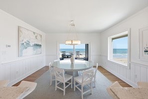Un coin repas avec une table en verre, des chaises blanches, un luminaire suspendu et une vue sur la plage par une grande fenêtre.