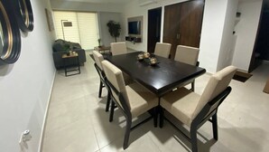 A modern dining area with a dark wooden table, beige upholstered chairs, and a television mounted on the wall. - Depas VAL704 By Trvl2hm Mazatlan Regency (Mazatlán)