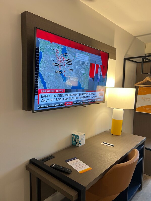 A hotel room with a TV mounted on the wall displaying a news channel, a desk with a lamp, a remote control, and a small box. - Comfort Inn (Princeton)