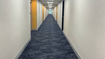 Un pasillo largo y estrecho con alfombra estampada, paredes blancas e iluminación fluorescente en el techo.
