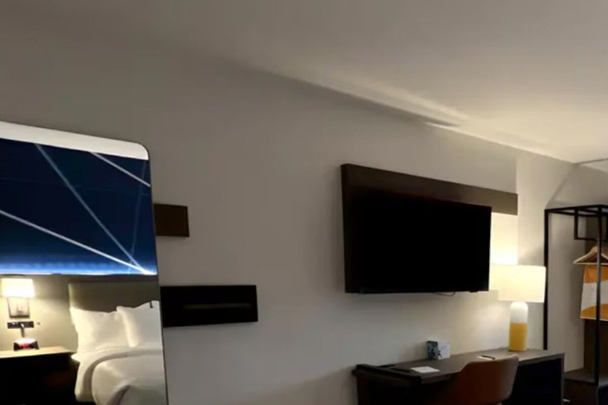 Ein Hotelzimmer mit einem Bett, einem Schreibtisch, einem Stuhl, einem Fernseher, einem Spiegel und einem Wandfernseher.