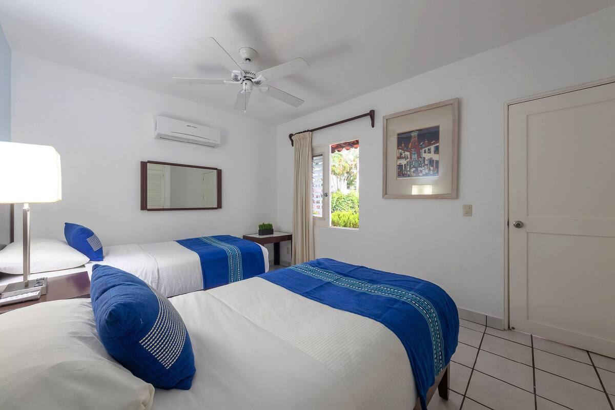 Habitación de hotel con dos camas, un ventilador de techo, un aire acondicionado, una ventana con cortinas, un cuadro enmarcado y una puerta.