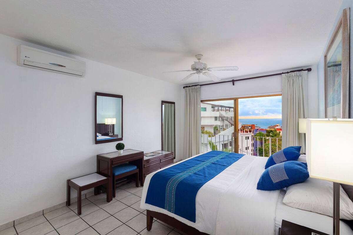 Habitación de hotel con cama, escritorio, silla, ventilador de techo y vistas a la ciudad.
