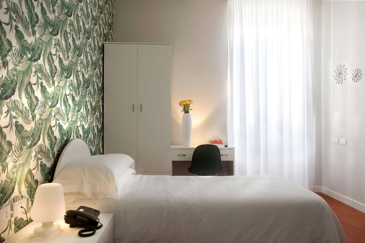 A hotel room with a bed, a desk with a chair, a lamp, a phone, a vase with flowers, and a wardrobe.