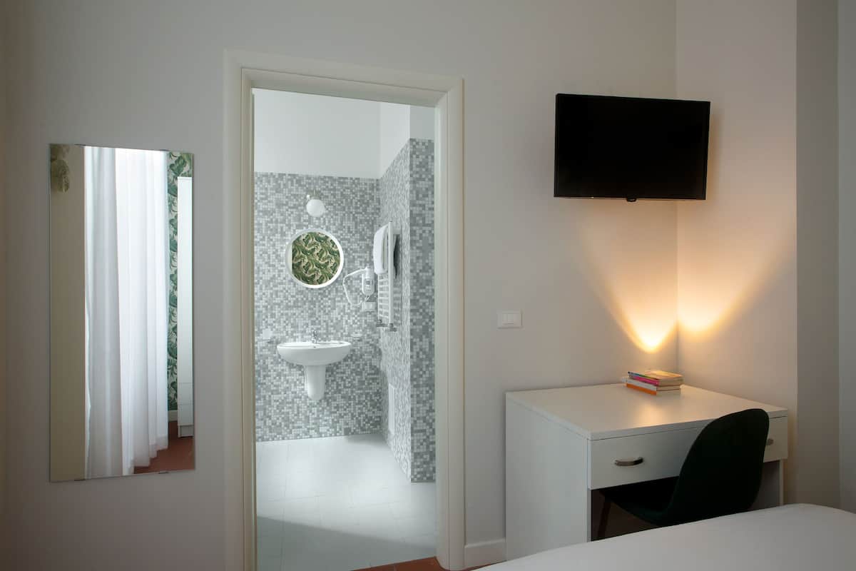 A hotel room with a bed, a desk with a chair, a TV mounted on the wall, and a bathroom visible through an open door.