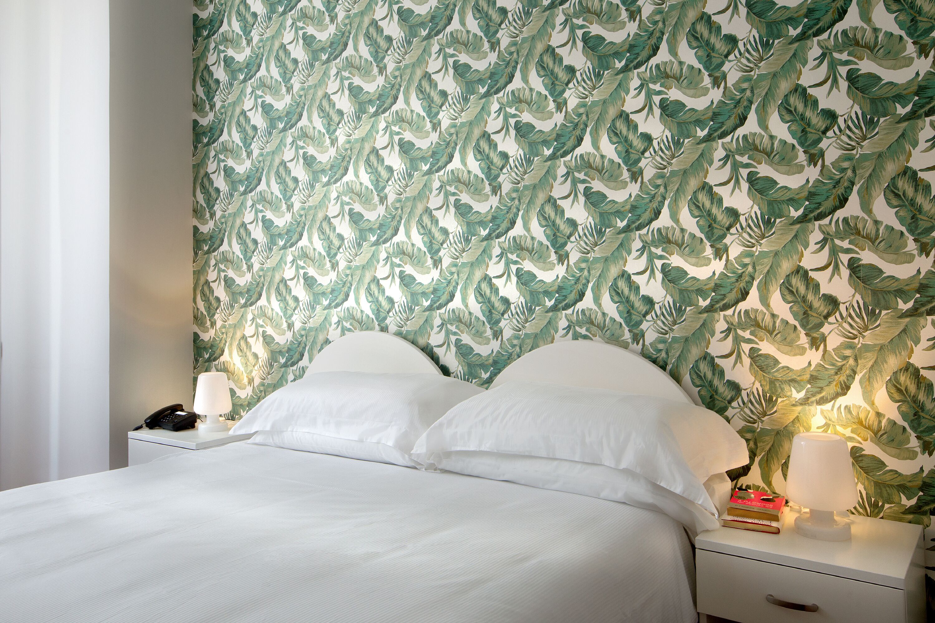 A neatly made bed with white linens, a bedside lamp, and a nightstand with a phone and books.