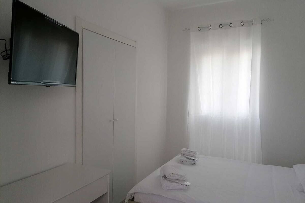 A bedroom with a bed, a TV mounted on the wall, a wardrobe, and a window with curtains.