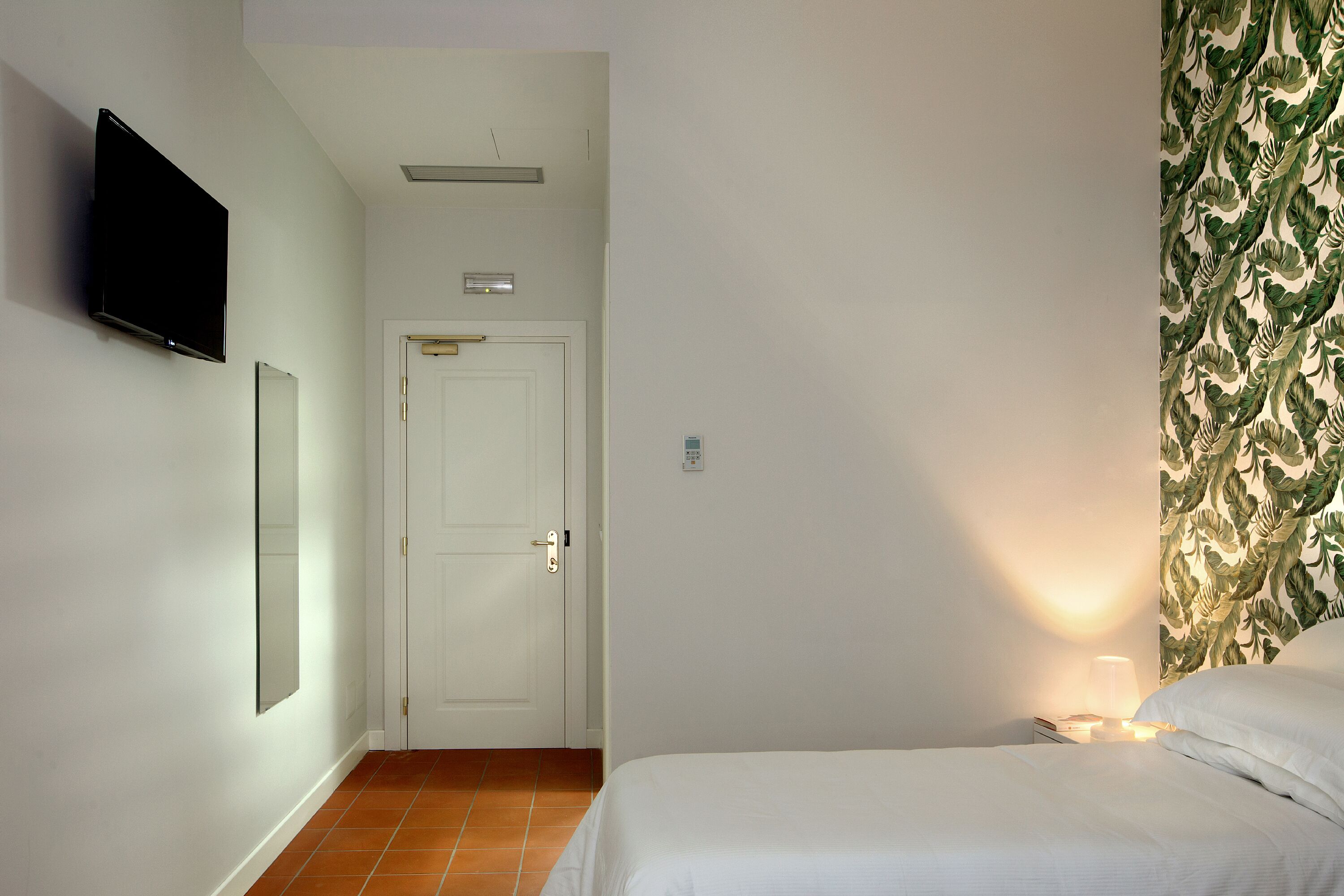 A bedroom with a bed, a nightstand, a wall-mounted TV, a door, and a patterned wallpaper.