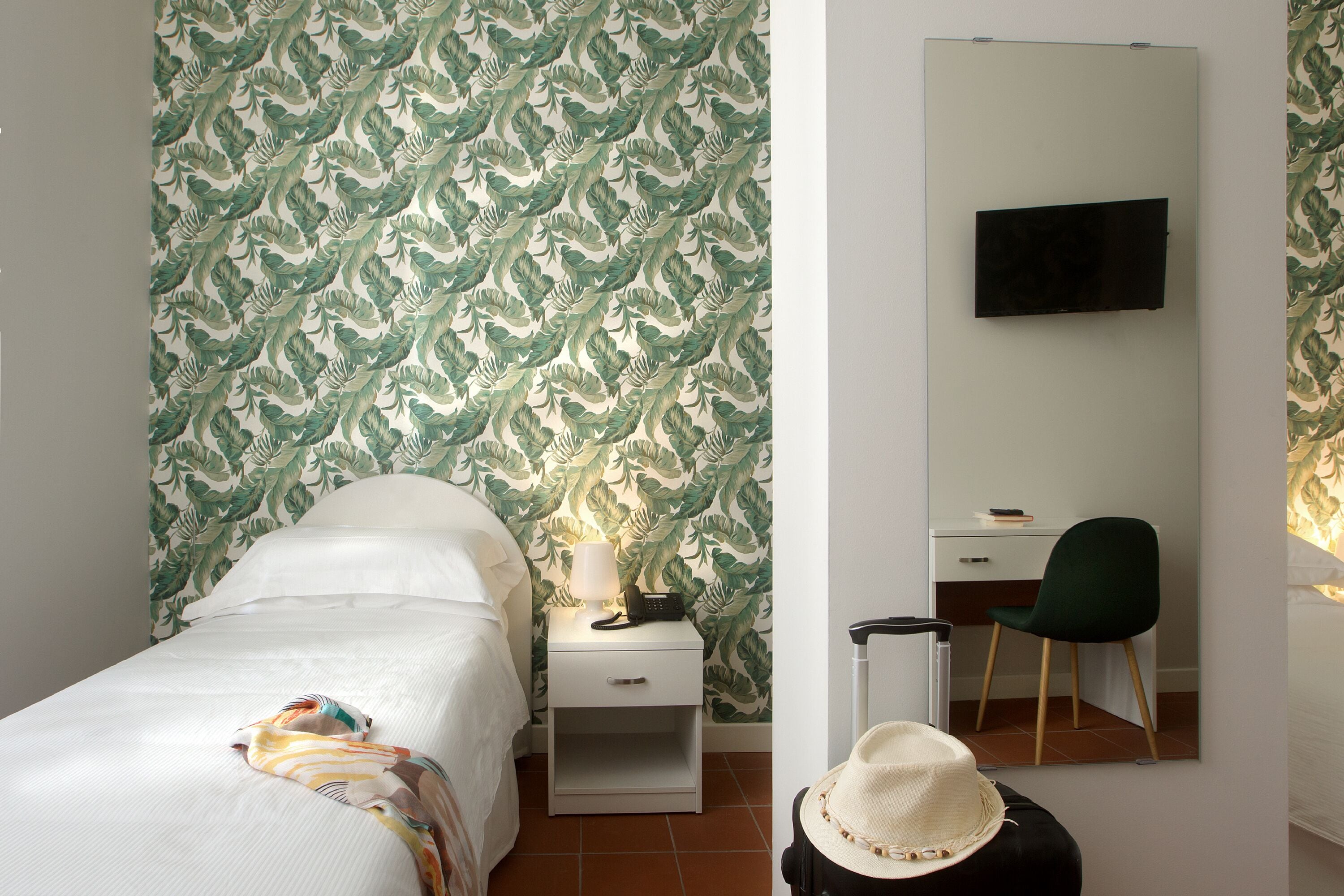 Habitación de hotel con cama, mesita de noche con lámpara, escritorio con silla y televisor montado en la pared.