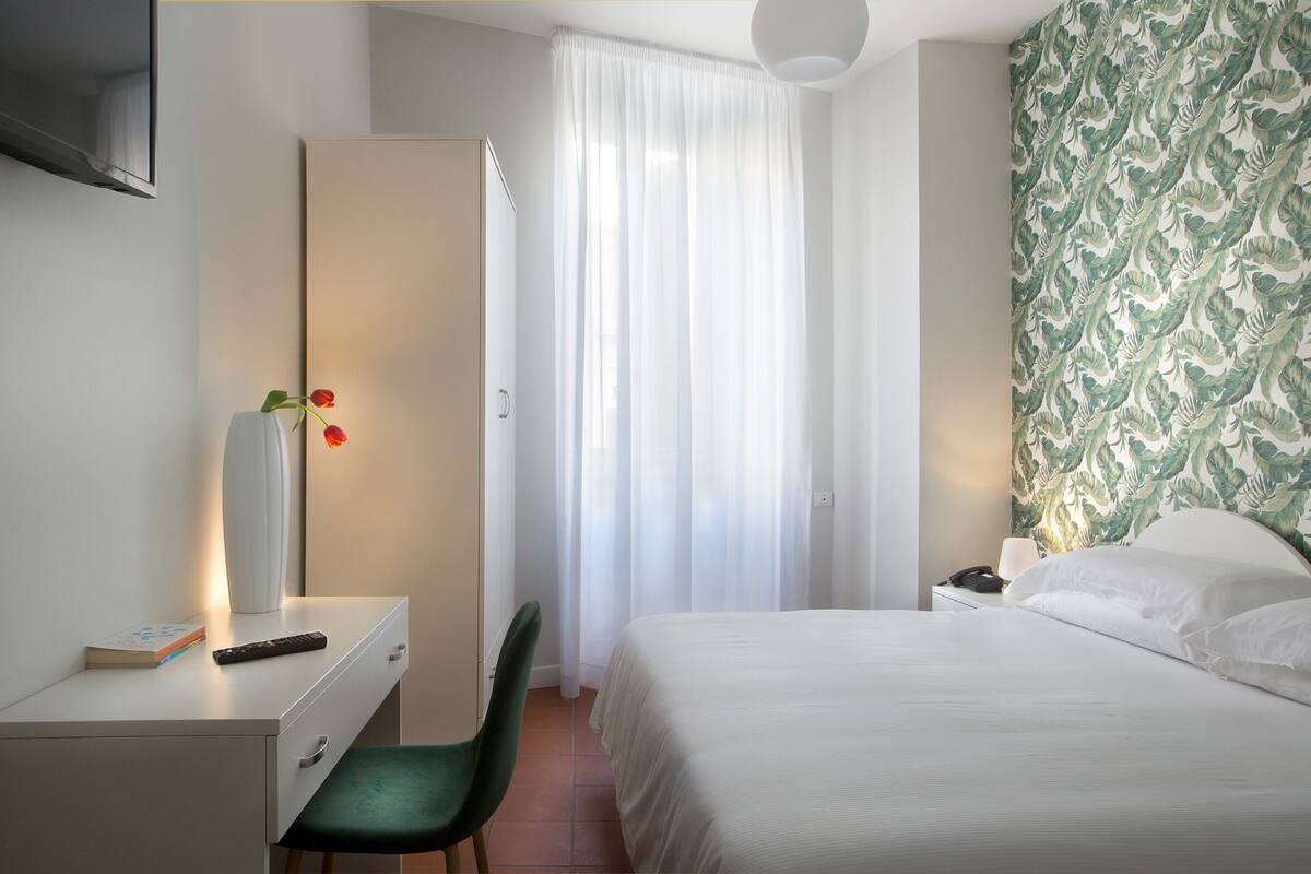 A hotel room with a bed, a desk with a chair, a bedside table with a lamp, and a curtain.