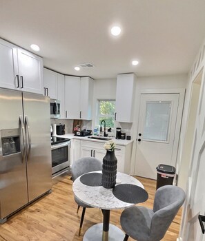 A modern kitchen with white cabinets, stainless steel appliances, and a marble-top dining table with grey chairs. - AIRPORT LOUNGE INN (College Park)