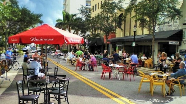 Un café al aire libre con mesas y sillas, una sombrilla roja de Budweiser, y personas comiendo.