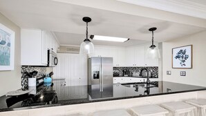 A modern kitchen with white cabinets, a stainless steel refrigerator, a black countertop, and bar stools. - Beachfront |Pool, Games, No Stairs! Sleeps 12 (Destin)