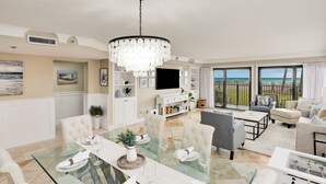 A spacious living room with a glass dining table, comfortable seating, a large chandelier, and a view of the ocean through large windows. - Beachfront |Pool, Games, No Stairs! Sleeps 12 (Destin)