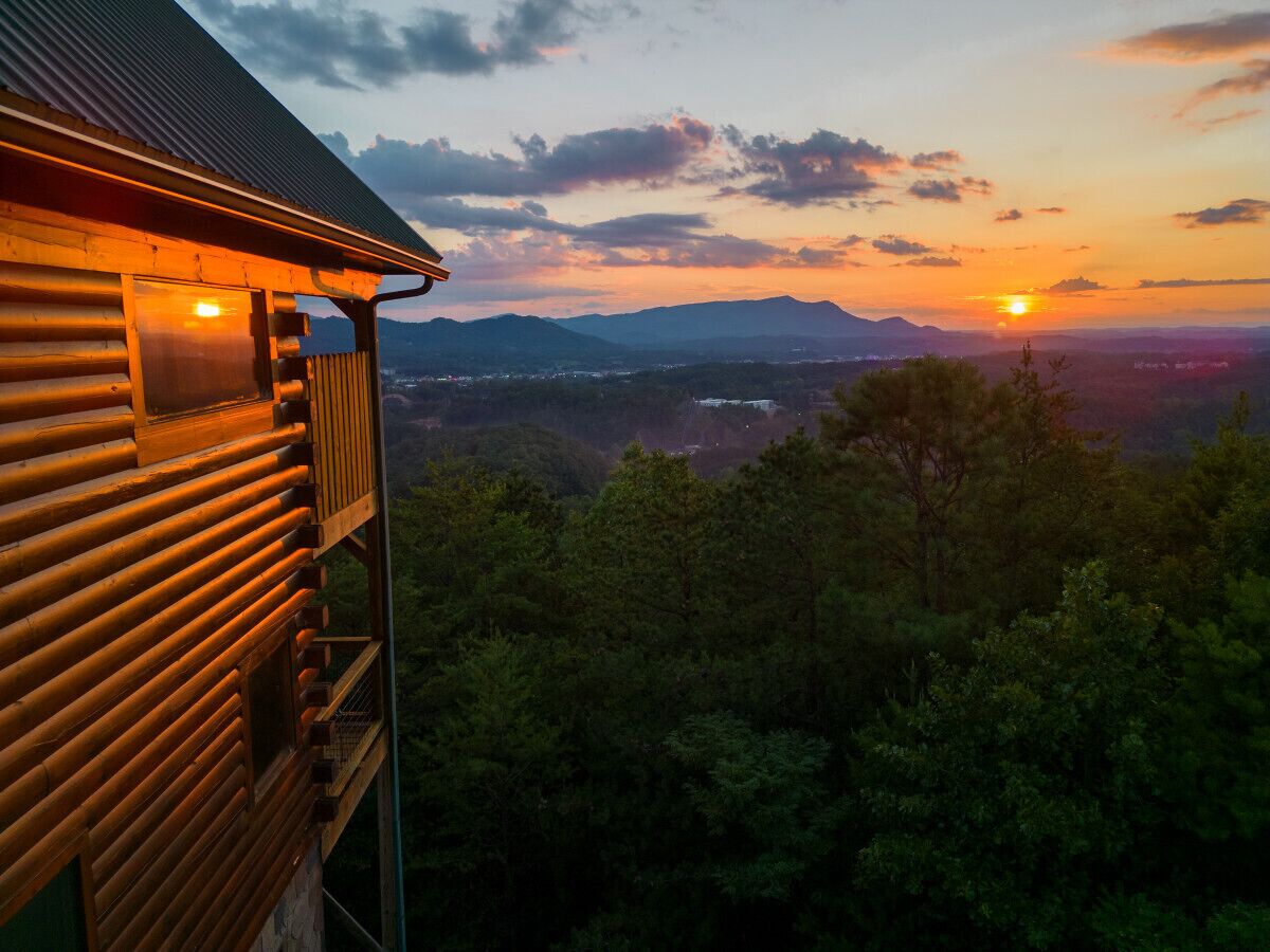 A wooden cabin with a balcony overlooking a forested landscape and a sunset in the background.