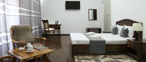 A hotel room with a bed, a desk, a chair, a TV, and a table with a teapot and book.