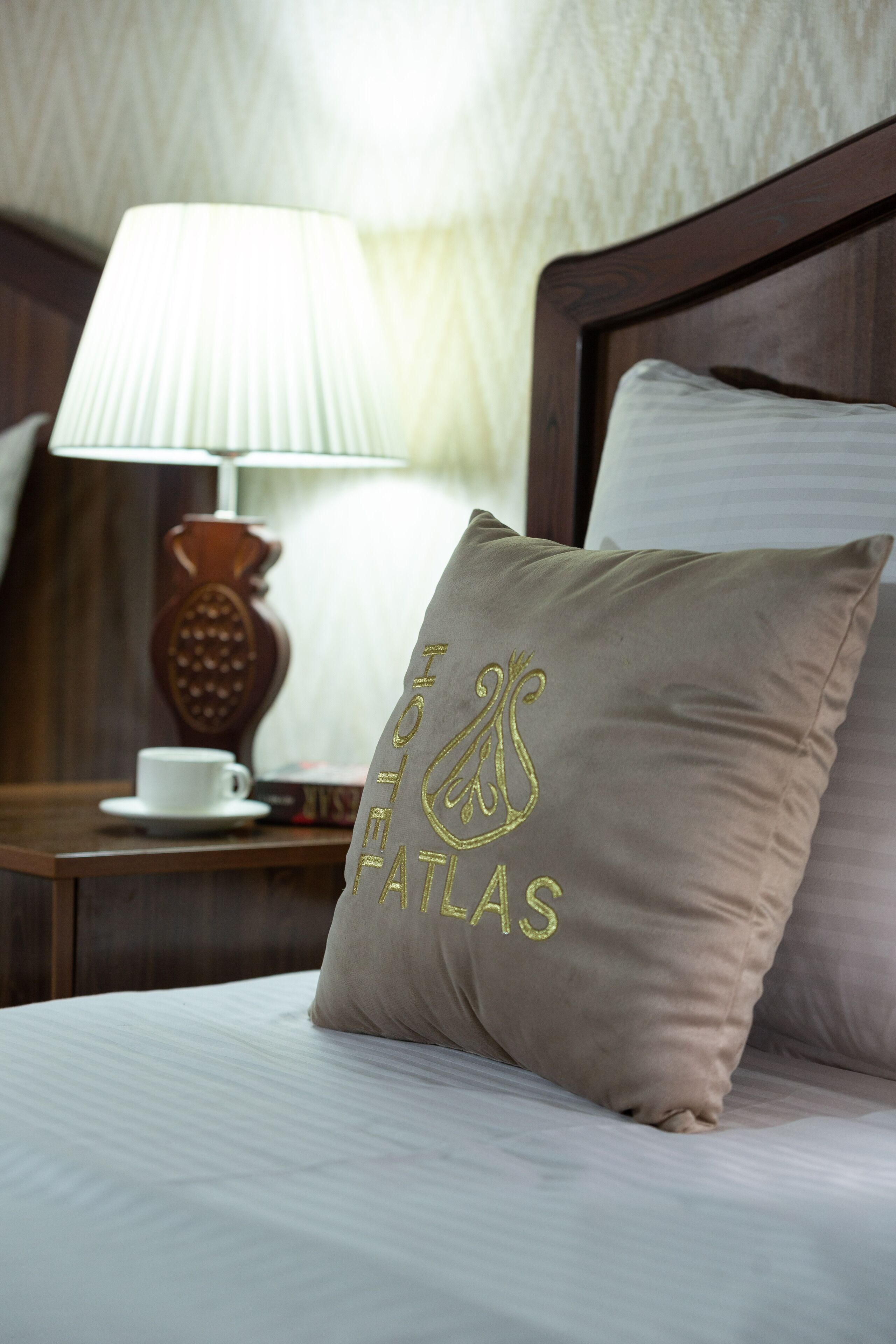 A neatly made bed with a pillow featuring embroidered text, a bedside table with a lamp, and a cup.