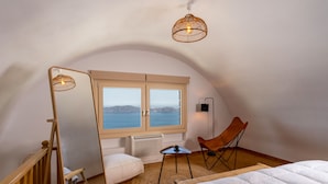 Un dormitorio acogedor con una cama grande, una silla, una mesita, un espejo y vista al mar a través de una ventana.