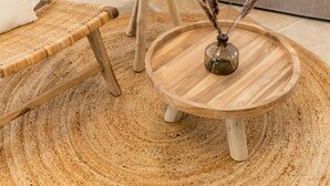 Una sala de estar con un sofá color beige, un sillón de mimbre, una mesita de madera y una mesa redonda de madera con un jarrón de plantas secas.