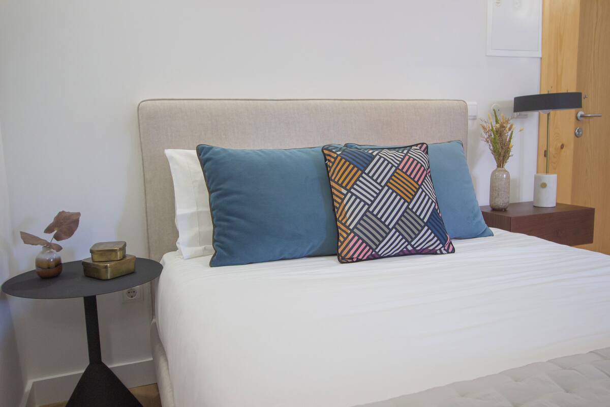 Una cama bien hecha con un cabecero color beige, dos almohadas y una mesita de noche con un florero y un adorno.