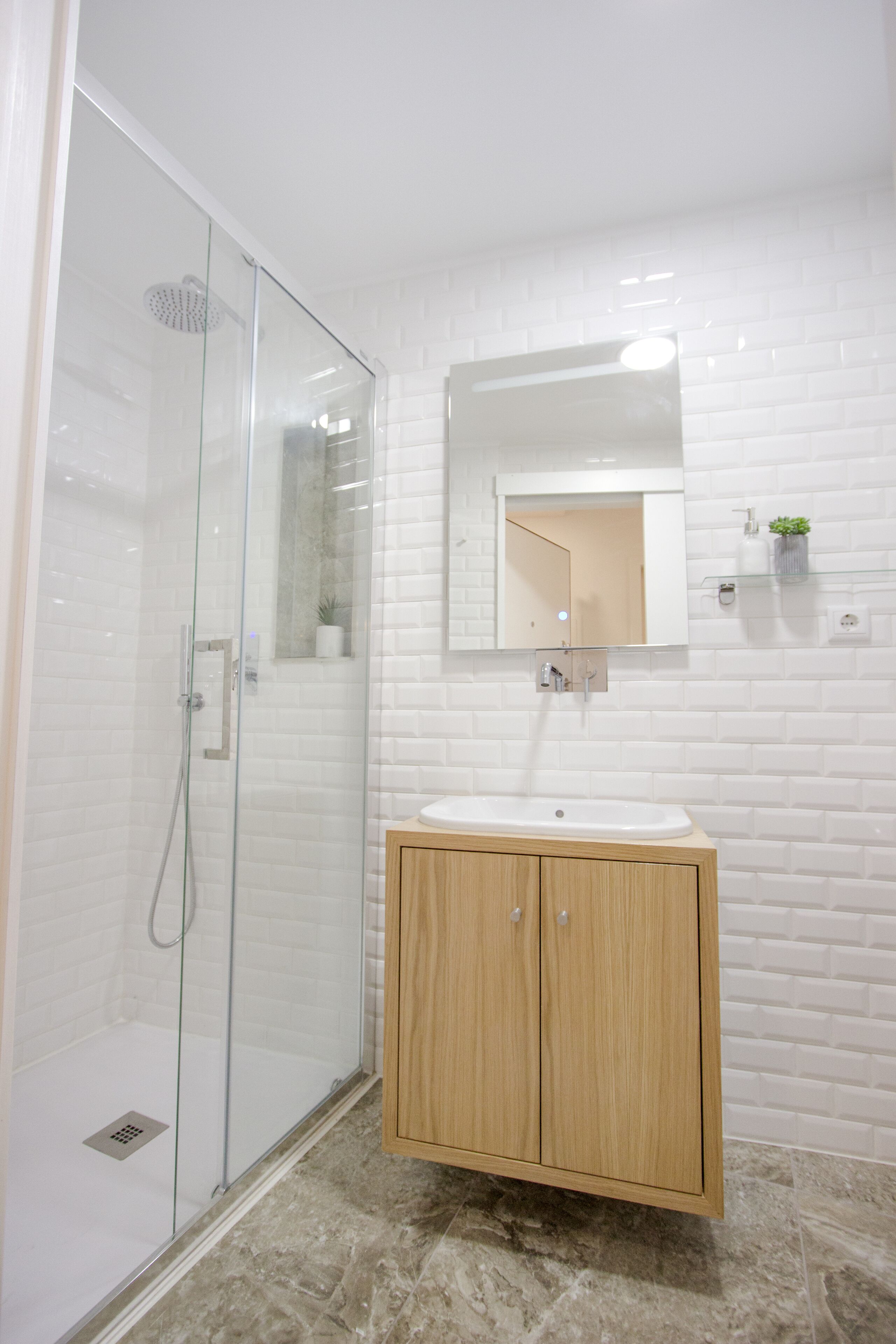 Een moderne badkamer met een glazen douchecabine, een houten wastafelmeubel met een wastafel en een betegelde muur in het wit.