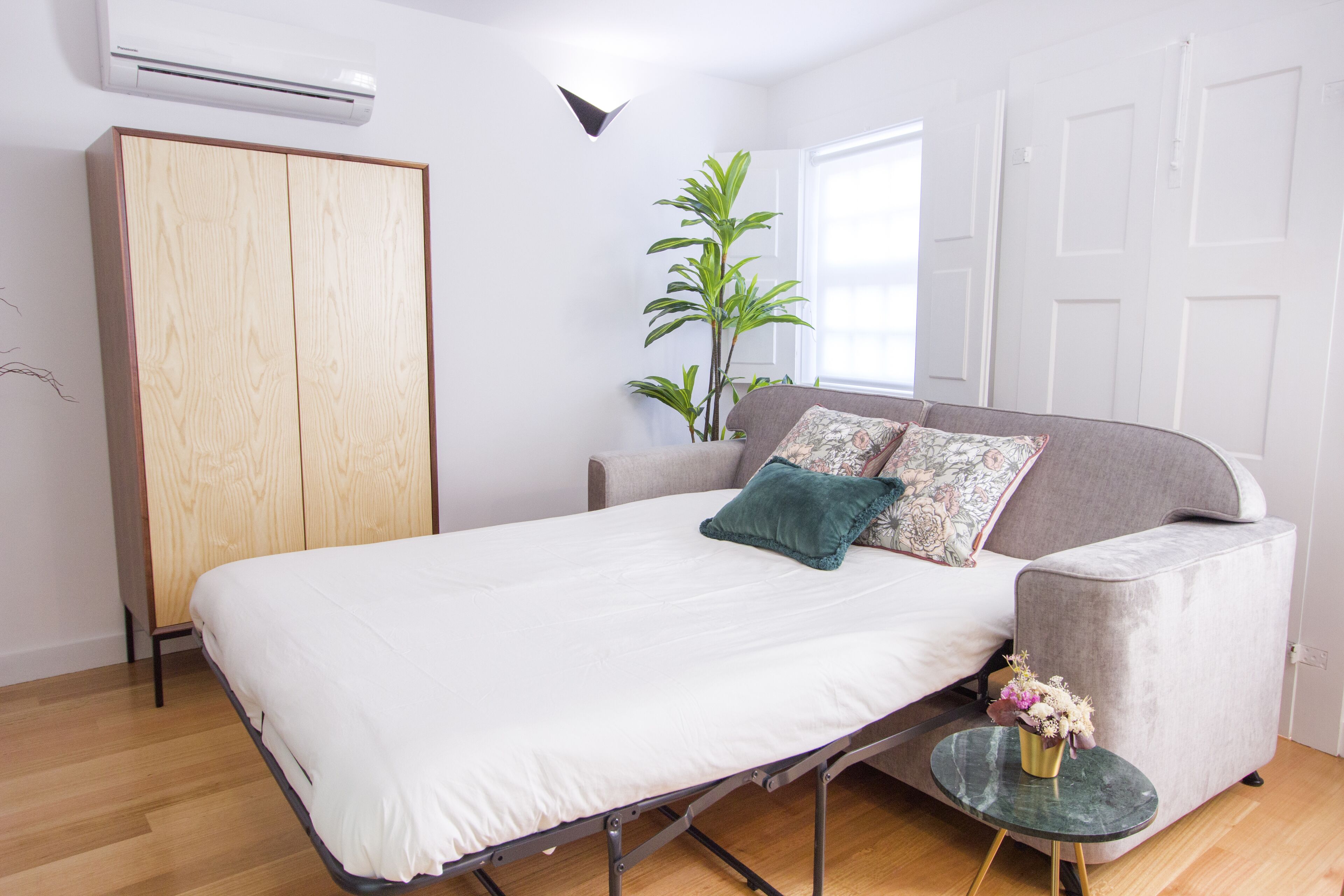 Ein Schlafzimmer mit einem Bett, einem grauen Sofa, einem kleinen runden Tisch mit einer Vase und einer hohen Pflanze.