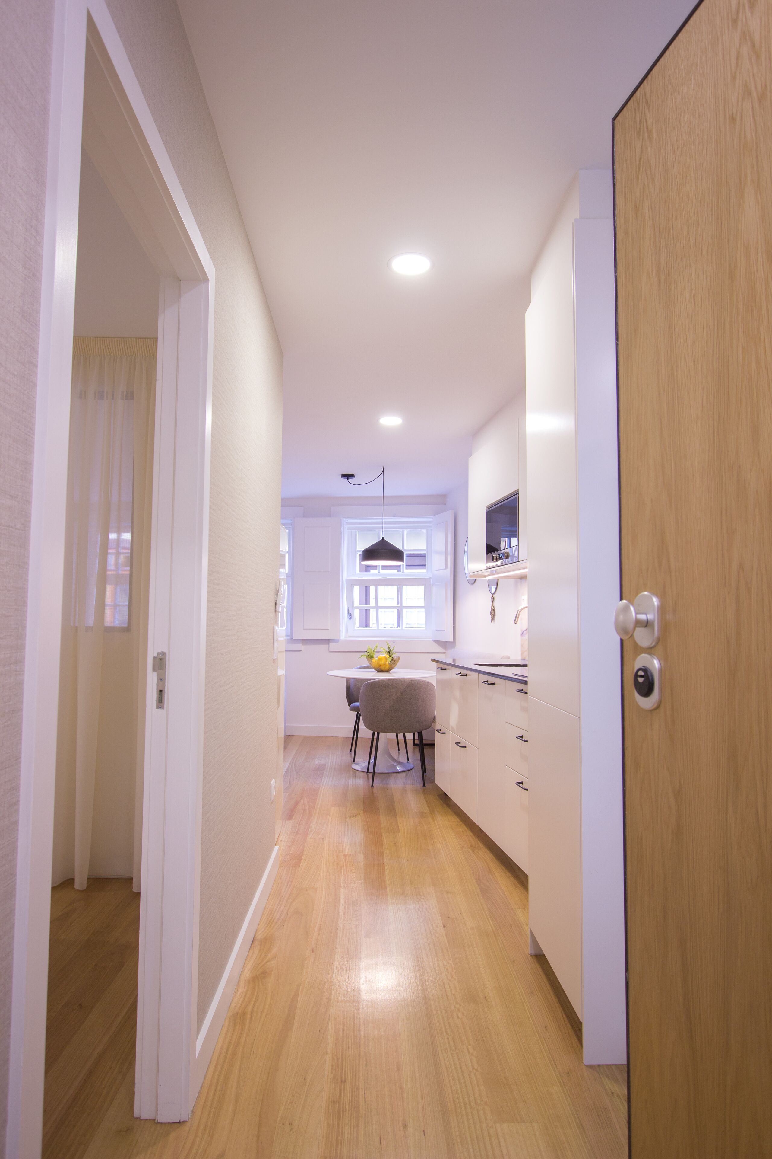 A narrow hallway with wooden flooring leading to a kitchen area with white cabinets and a window.