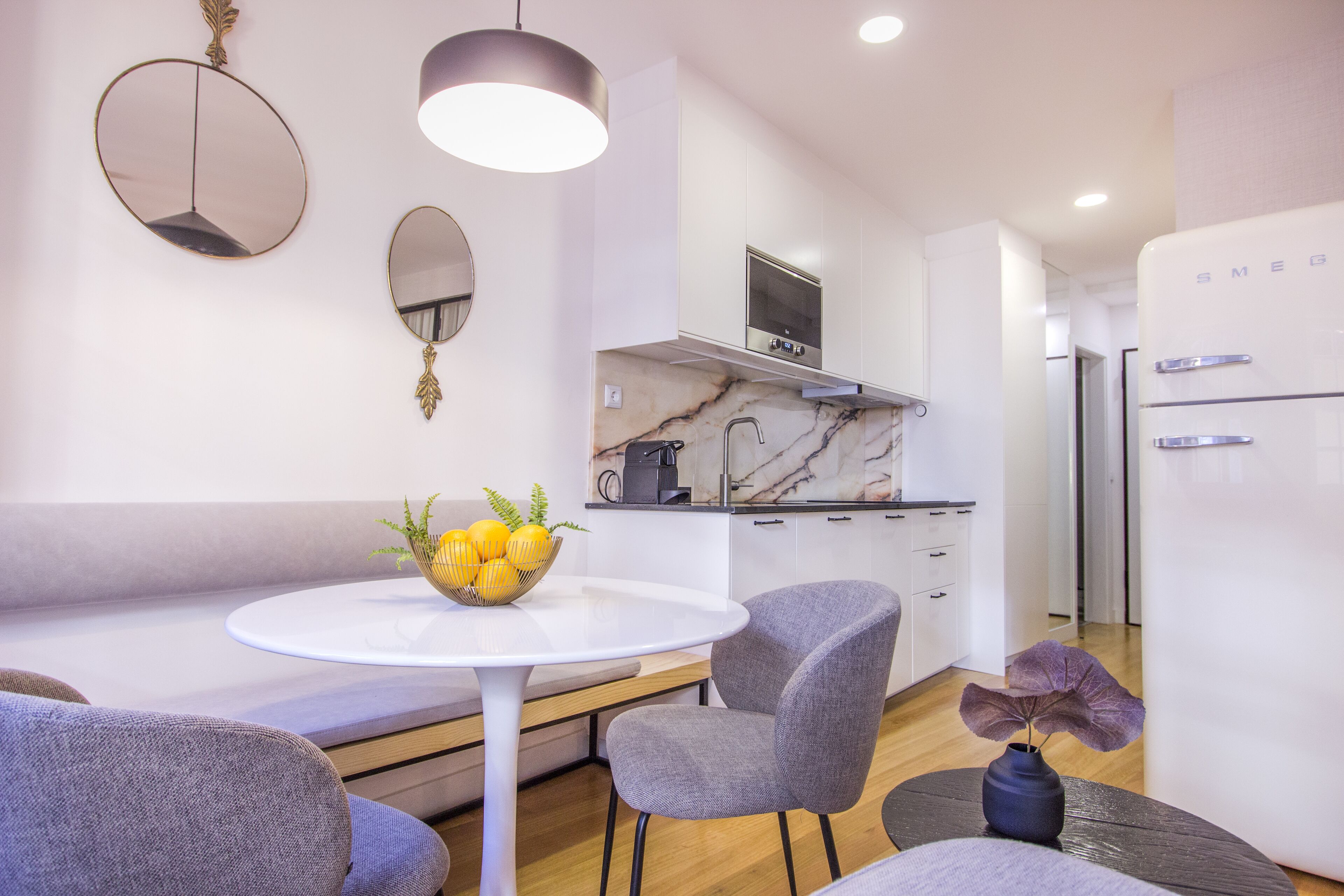 A modern kitchen with white cabinets, a marble countertop, and a built-in oven. There is a dining area with a round table and two chairs.
