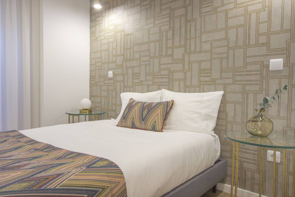 Una cama bien hecha con una colcha estampada, una almohada decorativa y una mesita de vidrio con un florero.