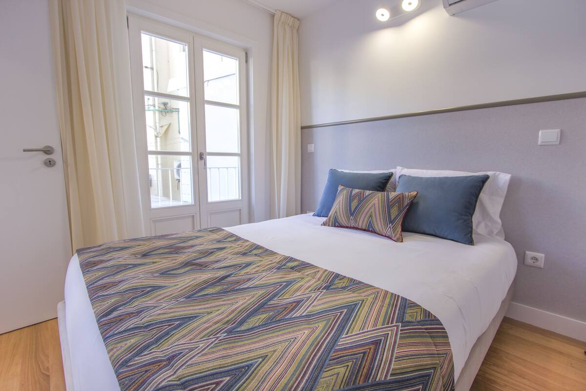 Una cama bien tendida con una colcha estampada, una ventana con cortinas y un aplique de pared.