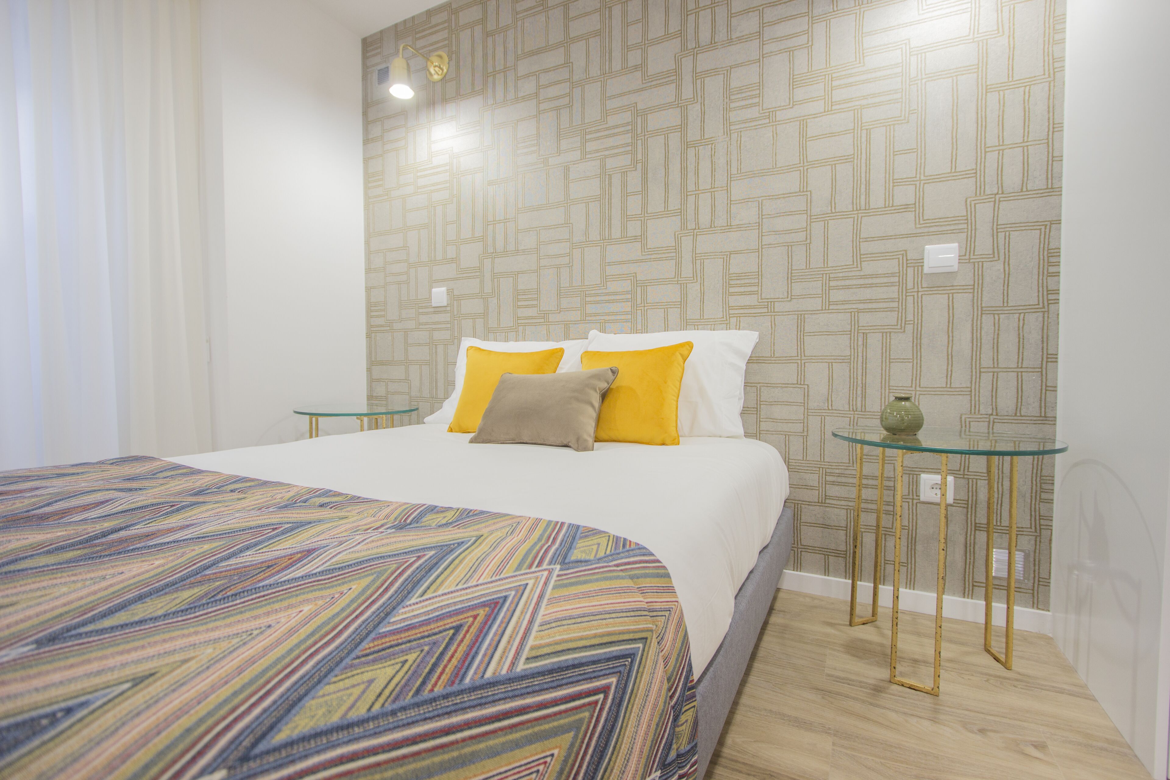 Un dormitorio ordenado con una colcha estampada, un diseño geométrico en la pared y una pequeña mesa con un florero.