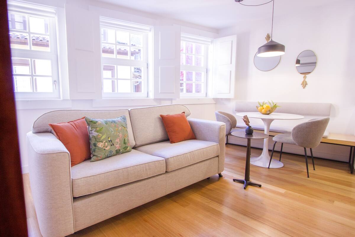 Una sala moderna con un sofá color beige, dos cojines anaranjados, una mesa redonda con sillas y un ventanal con contraventanas blancas.