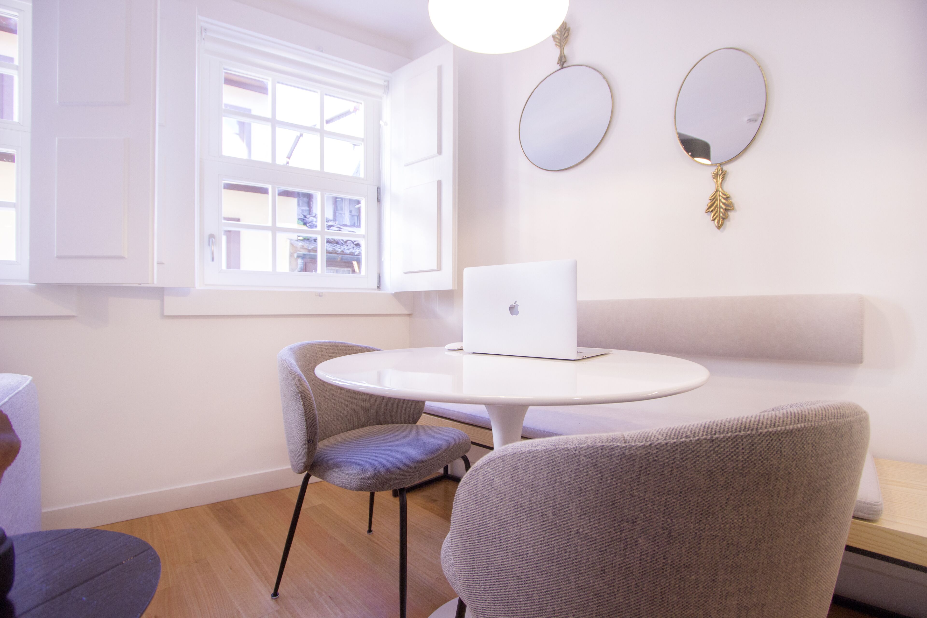 Una habitación con una mesa redonda, dos sillas, una computadora portátil, una ventana con contraventanas blancas y dos espejos colgados en la pared.