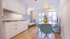 Una cocina moderna con armarios blancos, un comedor con mesa redonda y sillas azul verdosas, y un gran ventanal que proporciona luz natural.