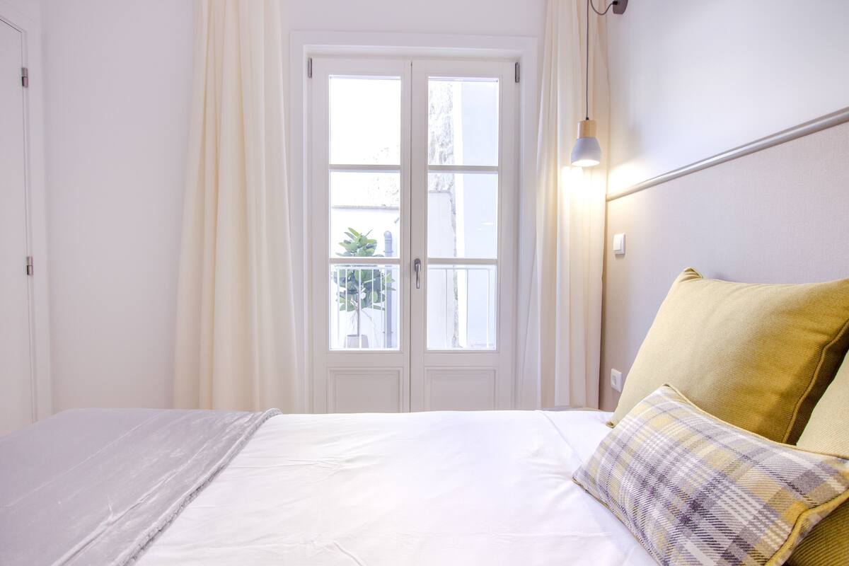 Un dormitorio con cama, cortinas, una ventana y una mesita de noche.