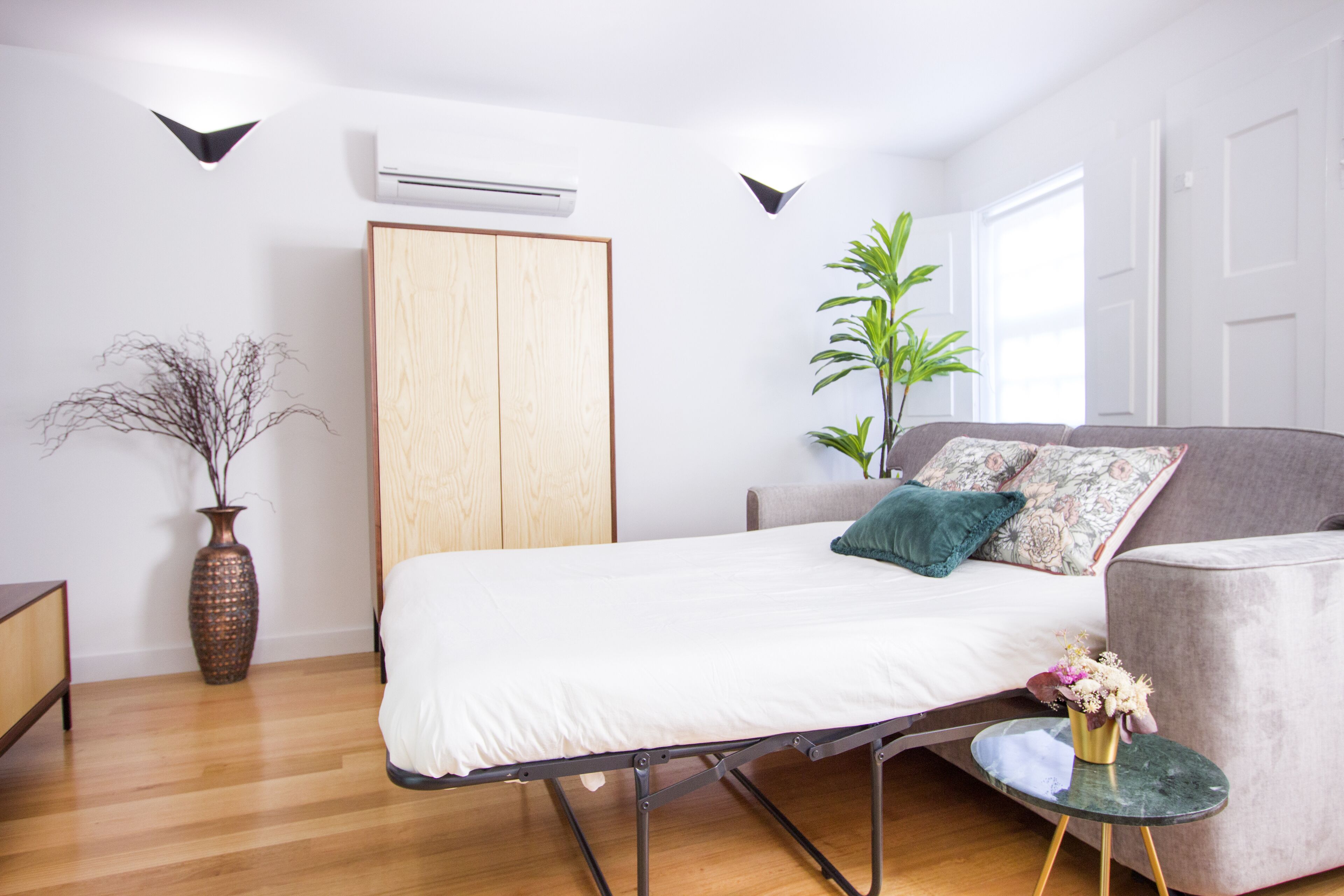 Une chambre à coucher moderne avec un lit, un canapé gris, une petite table avec un vase et une plante en pot.