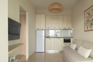 Cocina compacta con horno integrado, refrigerador y lavamanos. Hay un sofá color beige con cojines y un televisor de pantalla plana montado en la pared.