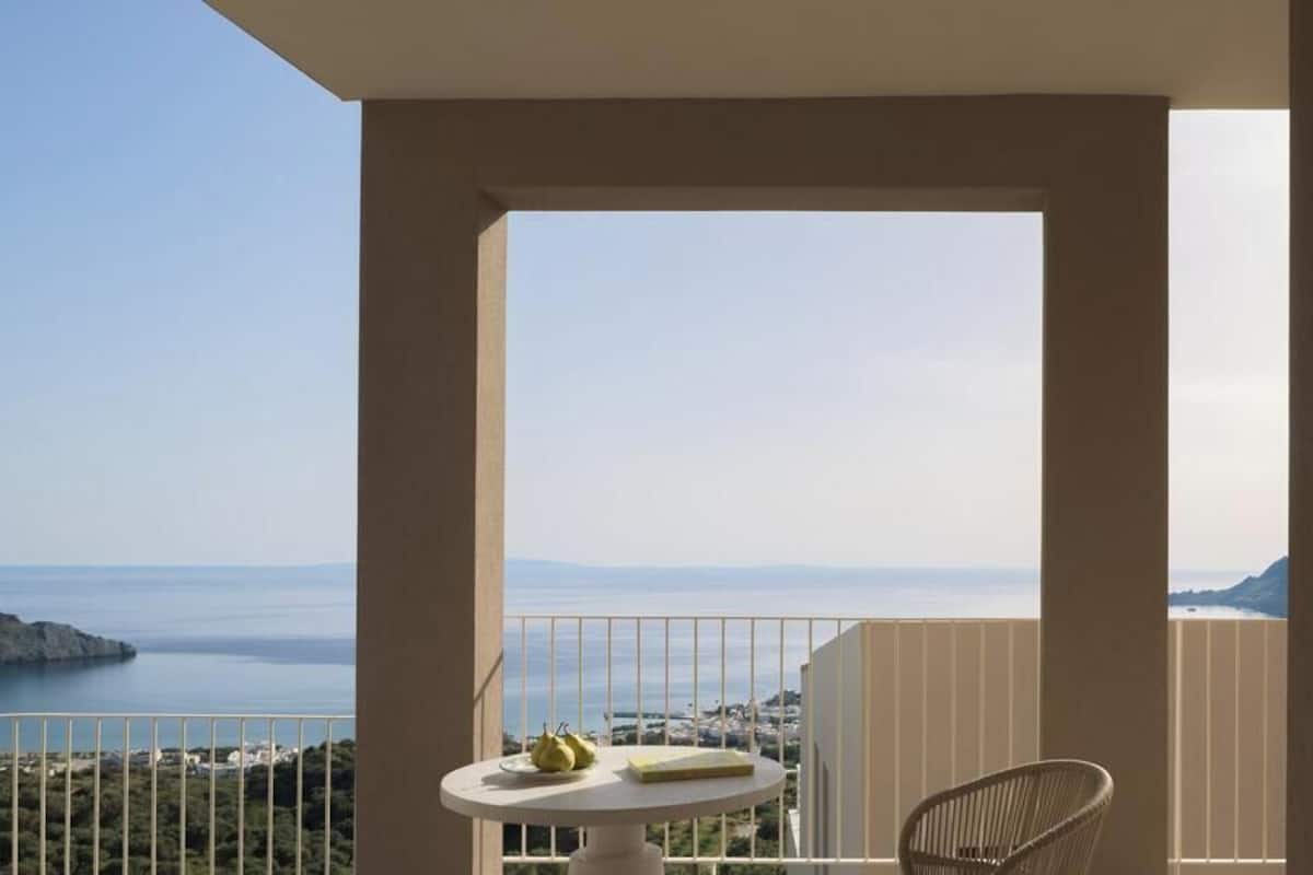 A balcony with a view of the sea, a table with fruit, and a chair.
