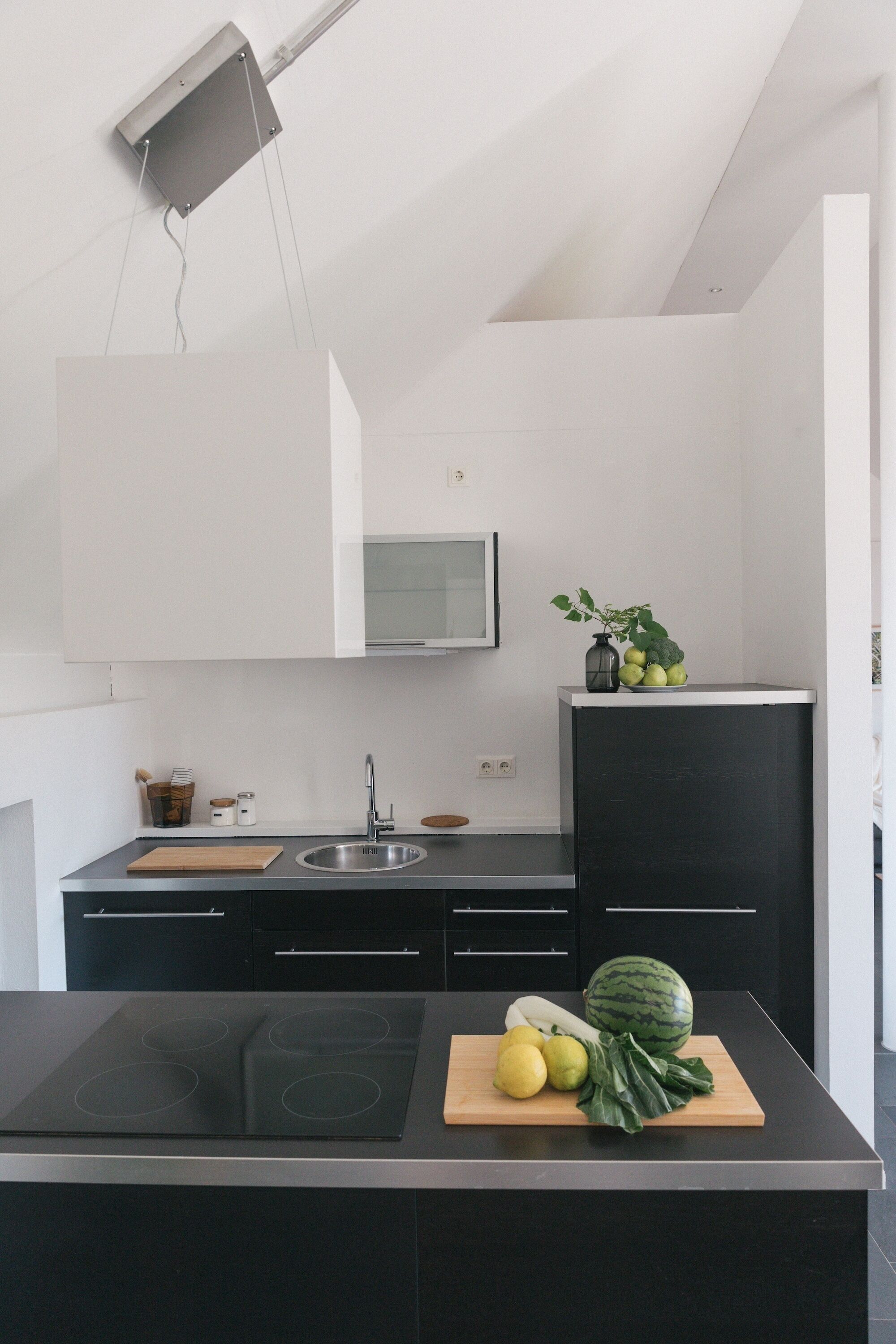 A modern kitchen with a black countertop, stainless steel sink, and a built-in stove. There is a wooden cutting board with vegetables and a potted plant on the countertop.