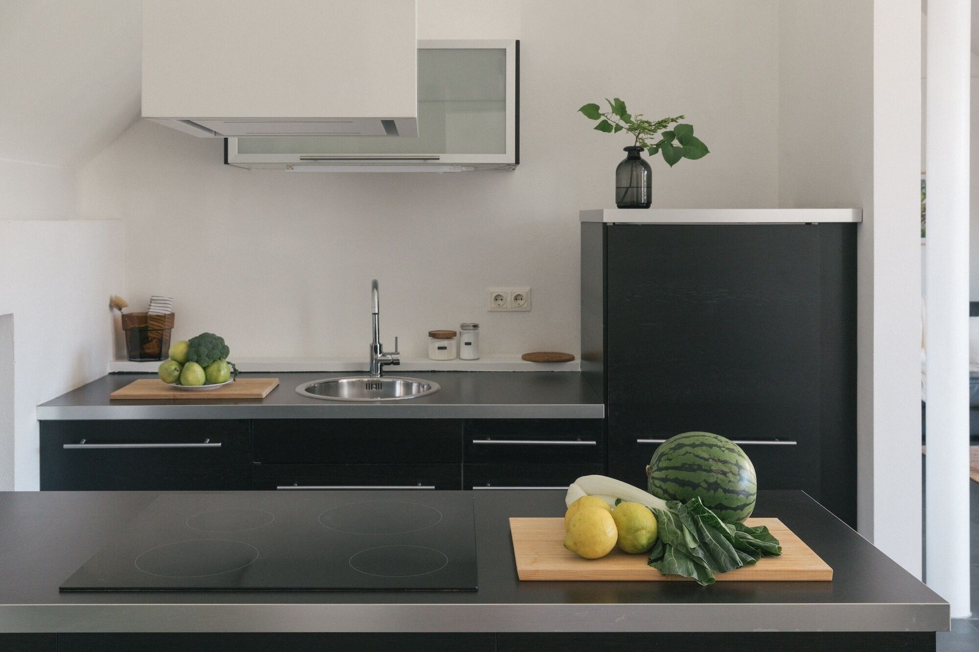A modern kitchen with a black countertop, stainless steel sink, and a black refrigerator. There are wooden cutting boards with fruits and vegetables on the counter.