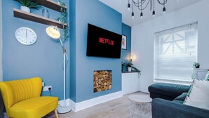 A living room with a yellow armchair, a dark sofa, a TV displaying 'NETFLIX', a clock, and a fireplace. - Stylish & Spacious home, Great Location, sleeps 6! (Chester)