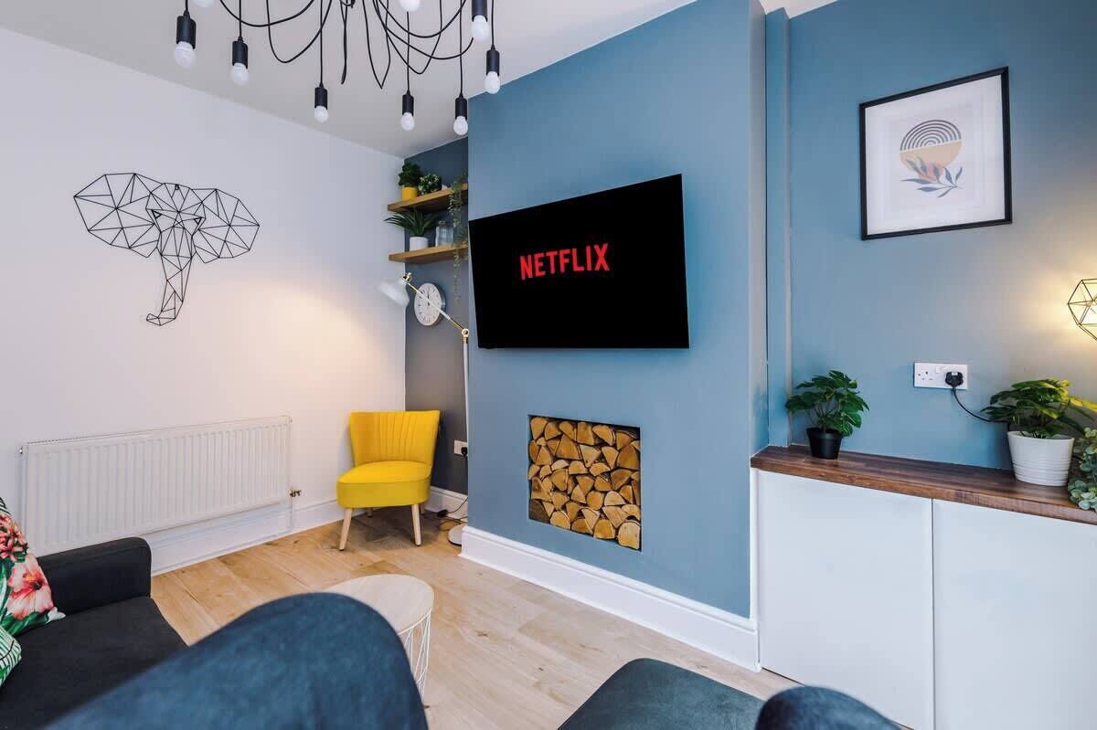 Uma sala de estar moderna com uma parede azul de destaque, uma TV fixada exibindo 'NETFLIX', arte abstrata na parede, uma poltrona amarela e um armário de madeira com plantas.