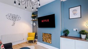 A modern living room with a blue accent wall, a mounted TV displaying 'NETFLIX', a geometric wall art, a yellow chair, and a wooden cabinet with plants. - Stylish & Spacious home, Great Location, sleeps 6! (Chester)