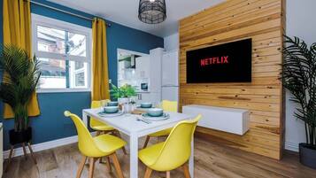 Una sala de estar moderna con una mesa de comedor blanca, sillas amarillas y un televisor montado en una pared de madera mostrando "NETFLIX".