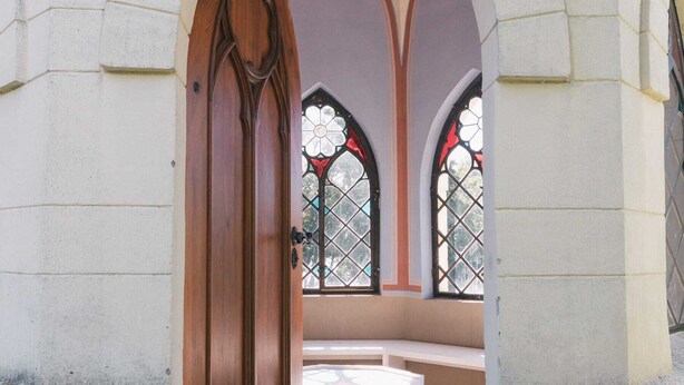 Un arc en pierre mène à un espace agrémenté d’un banc, avec une porte en bois et des vitraux.
