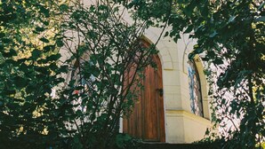 Une église dotée d’une porte en bois remarquable et de fenêtres en arc de cercle, entourée d’un feuillage dense.