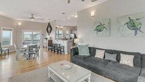 Ein geräumiges Wohnzimmer mit grauem Sofa, einem Couchtisch und einem Essbereich mit Tisch und Stühlen. Es gibt eine Küchenzeile mit Bar und Deckenventilator.