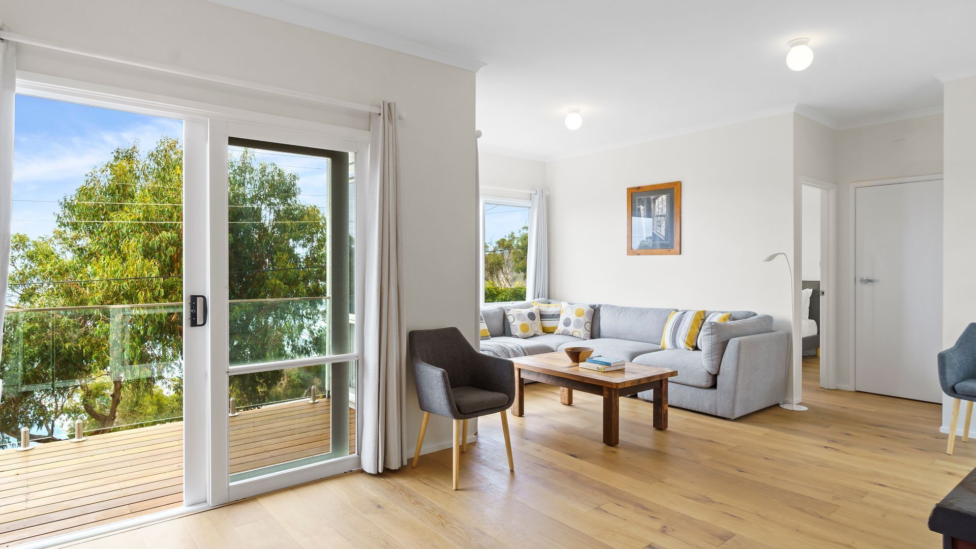 Een moderne woonkamer met een grijze bank, een houten salontafel en een zwarte fauteuil. Er is een schuifdeur die leidt naar een balkon met een houten terras.