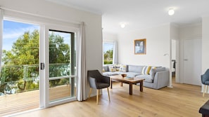 Un salon moderne avec un canapé gris, une table basse en bois et un fauteuil noir. Une porte-fenêtre coulissante donne accès à un balcon doté d’une terrasse en bois.